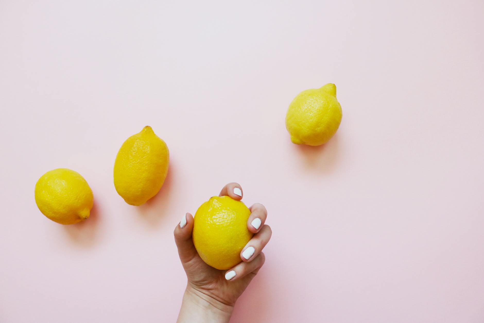 Hand holding a fresh lemon against a soft pink background with yellow lemons surrounding it.