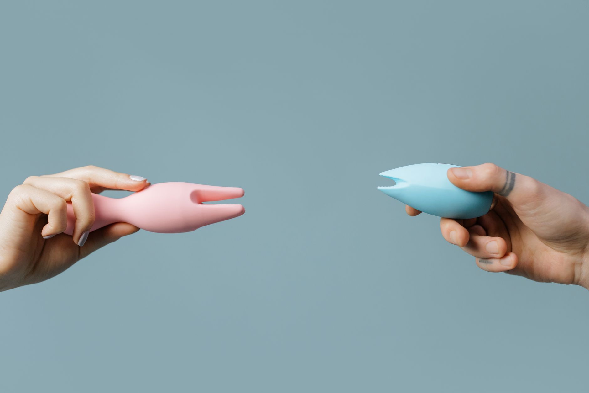 Two hands holding colorful silicone vibrators against a pastel background, representing openness and connection
